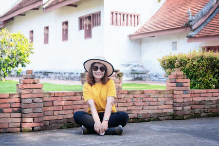 Portrait of adult beautiful Asian woman wearing a straw hat and sunglasses sitting on the floor with a temple background. Happy lady smiling and relaxing on summer vacation.の写真素材