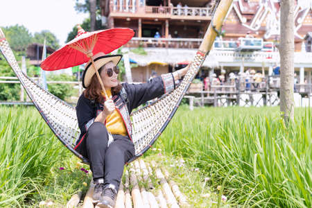 A cute adult Asian woman wears a straw hat and sunglasses holding a red paper umbrella handmade with green rice plants around and blurred tourist background on holiday during the coronavirus pandemic.の写真素材
