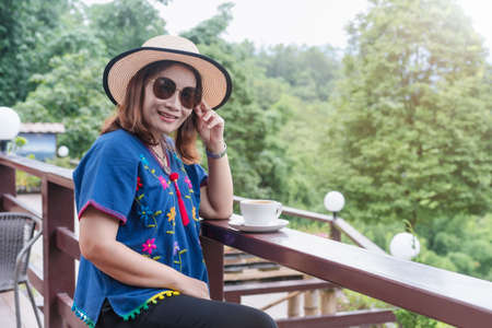 Beautiful adult Asian woman wearing a straw hat, sunglasses and blue native clothes of North Thailand smiling and relaxing with hot coffee mug in vacation sunrise and blurred green nature background.の写真素材