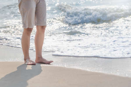 Young woman's bare feet with a wave coming and enjoying traveling to exotic nature. Beautiful healthy female legs with droplets standing barefoot alone on sandy sea beach in summer. Lifestyle conceptの写真素材