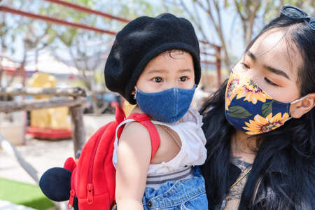 Little child girl and Asian mother wearing medical protective mask and holding her daughter in a crowded public place in park or tourist. Happy baby and mom family playing outside   pandemic.の写真素材