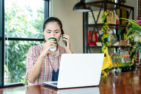 Young charming woman talking to the phone, working on a laptop and drinking coffee in a cafe.  Attractive female calling with her cell telephone. Student learning online. Freelancer working online.の写真素材