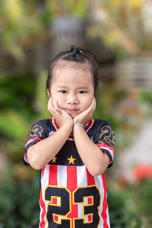 Portrait of adorable child girl looking at camera and gentle smile holding hands under chin looking contented and carefree. Cheerful female Kindergarten student posing against blurred green backgroundの写真素材