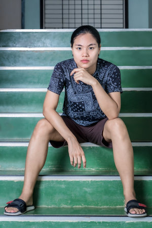 Portrait of confident young woman sitting on stairs and looking at camera. Beautiful Asian young female indoors sitting on steps, relaxing and thinking with hopeful and fighting problems conceptの写真素材