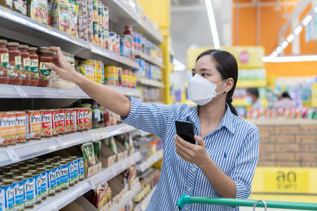Suphan Buri, Thailand-July 3, 2021: Young woman wearing medical face mask using smartphone to reading shopping list for choosing ketchup or food while shopping in supermarket during Covid-19 outbreakのeditorial素材
