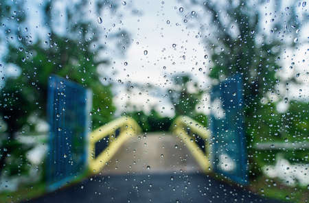 Rain droplets on the surface of car glass with blurred green nature and bridge background through window glass of car covered by raindrops. Freshness after rain. Wet windscreen shot from inside car.の写真素材