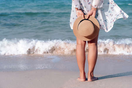 Back view of woman's legs standing on sandy beach near turquoise colored ocean in summertime. Female tourist with straw hat in hands enjoying traveling to exotic nature on a beautiful sunny day.の写真素材