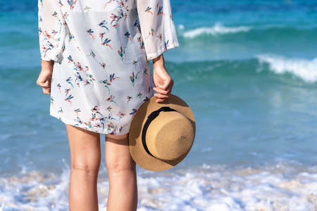 Back view of woman's legs standing on sandy beach near turquoise colored ocean in summertime. Female tourist with straw hat in hands enjoying traveling to exotic nature on a beautiful sunny day.の写真素材