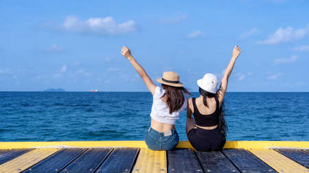 Back view of two happy young women rising hands up and sitting on  bridge with tropical ocean...
