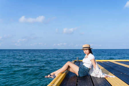 Worried woman sitting on a bridge with a straw hat tropical ocean by the sea in a sunny day time. Lonely girl sitting alone on the seaside, relaxing and thinking. Human emotion concept photographyの写真素材
