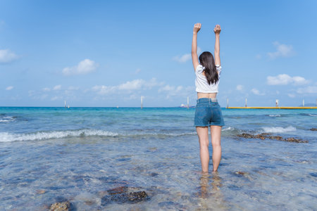 Happy young woman rising hands up and standing on beach with tropical ocean backgrounds, enjoying the beautiful view of the blue sea. Outdoor summer lifestyle. Vacations and freedom concept.の写真素材