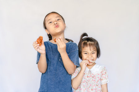 Close up portrait of two satisfied adorable Asian girls eating fried chicken. Happy small hungry kids biting fried chicken and enjoying a meal together. Children's healthy diet. Unhealthy food conceptの写真素材