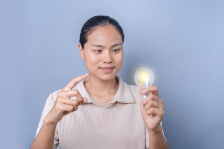 A young Asian woman points her finger at a glowing light bulb in her hand, having a creative idea, inspired by the light bulb. Innovation and electric energy concept. on a gray background.の写真素材