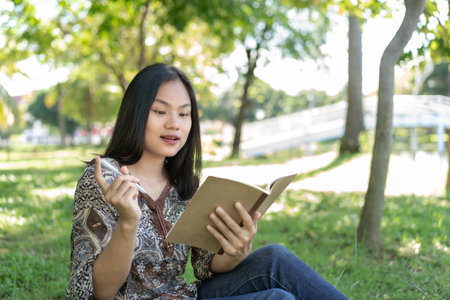 Asian teenage girl Wearing traditional clothes sitting on the grass holding a book and pen in hand happily a diary of daily life stories, planning ahead, ideas, and inspiration.の写真素材