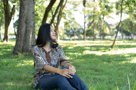A young Asian woman sits on the grass with her knees hugged, looking around. Relax, feel free,...
