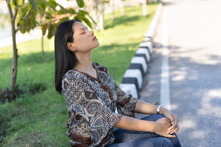 Attractive Asian woman sits with her arms crossed on her knees, eyes closed, face up, feeling content, calm, breathing in the cool, fresh air. While waiting for the bus on the side of the road.の写真素材