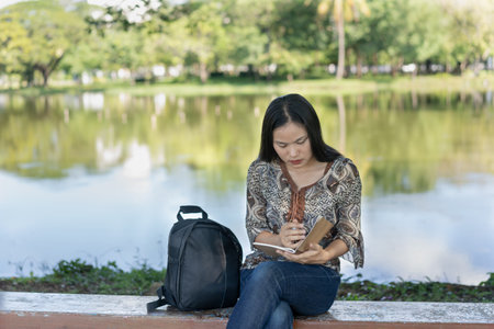Beautiful young woman, sitting on a stone chair, looking down at a notebook, holding a pen Record stories, keep a diary, black backpack beside you, quiet background, lake in the park.の写真素材