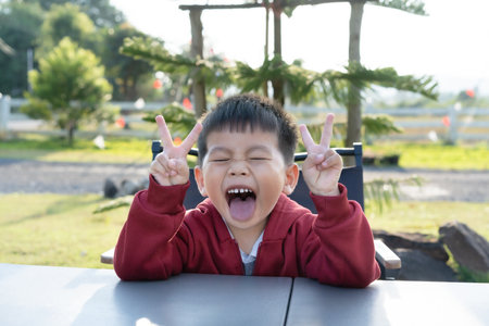 A cheerful little boy wearing a red sweater, smiling, in a good mood, sticking out his tongue, eyes closed, cheeky expression, and showing a two-fingered peace sign. Encouragement, friendliness.の写真素材