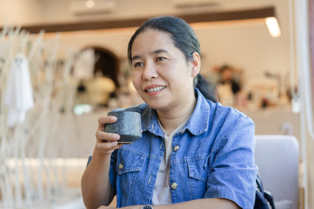 Happy woman wearing a blue jeans jacket sits holding a coffee cup. Looking forward to enjoying drinking in a cafe feeling of relaxation Concepts about business and happiness.の写真素材