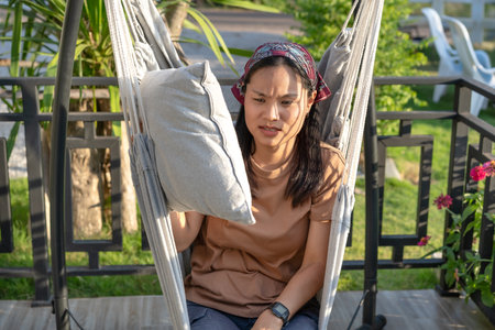Close-up of a young Asian woman lounging on a hammock outside her house with sunlight shining on her face. Feeling irritated, unhappy, bored, living at home alone, lonely concept.の写真素材
