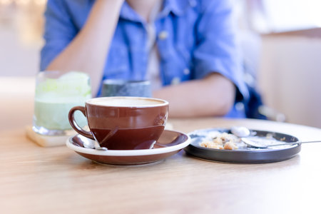 Focus on coffee mug and empty plate with bits of pastry and spoon in the plate. placed on the table On a blurry background, a woman sits with her hands on her chin. In the cafe, the atmosphere is warmの写真素材