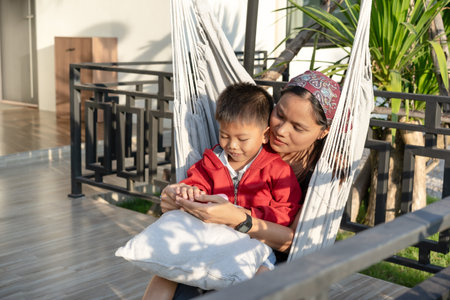 Happy moment, Asian mother sitting in a hammock with her little son, teasing, having fun, mother-child bonding, outside the house, sunlight shining, warm, during the weekend.の写真素材
