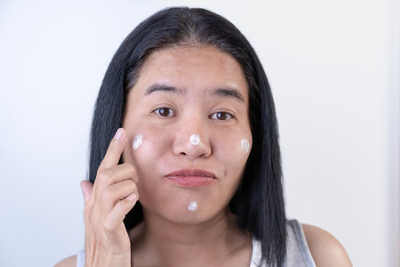 The playful woman uses her index finger to gently rub the cream into her cheek focused on maintaining her skin's health and hydration skincare application indicating a routine step in moisturizing.の写真素材