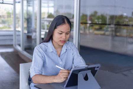 Young Asian woman working on tablet with stylus, focused and productive in a modern indoor/outdoor setting. Ideal for themes of remote work, technology, and casual businessの写真素材