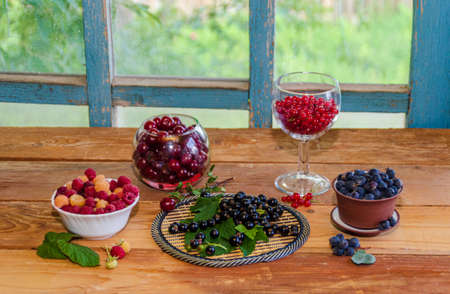 assorted berries on the table. Raspberries, cherries, irga, black and red currants with green leaves in a glass dish on a wooden table near the window. High quality photoの写真素材
