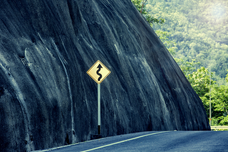 Yellow winding road sign at roadside beside hill stone.の写真素材