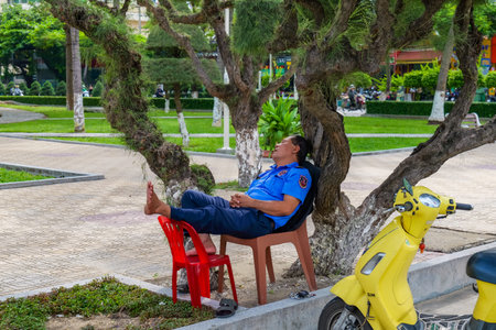 Uniformed man sleeping under a tree in an urban park, taking a comfortable break with his feet up on chairs, next to a yellow scooter. Nha Trang, 26.11.2025, Vietnamのeditorial素材