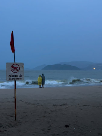 Two people in raincoats stand by the stormy sea near a warning sign in Nha Trang, 06.11.2025, Vietnam.のeditorial素材