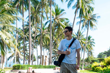 Young Man in White T-Shirt and Sunglasses Holding Iced Coffee Walking through Tropical Palm Tree Park in Bright Sunlight with Cinematic Depth of Field Nha Trang 20.12.2025 Vietnamのeditorial素材