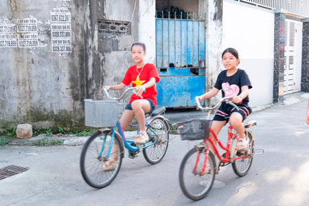 Two young girls ride bicycles along a paved alleyway past a textured wall. One wears a red shirt with a yellow star while the other is in black attire Nha Trang 20.12.2025 Vietnamのeditorial素材