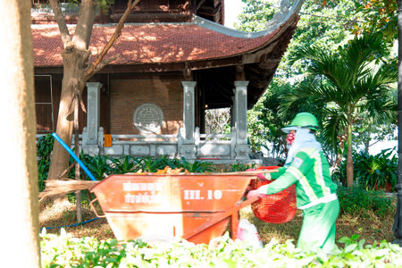 Asian Sanitation Worker in Green Uniform Pushing Waste Cart Near Traditional Pagoda, Cinematic Sunlight and Motion Blur, Environmental Service Concept Nha Trang 20.12.2025 Vietnamのeditorial素材