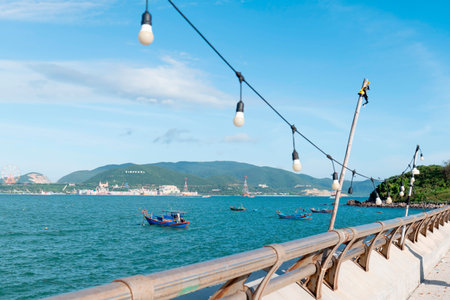 A daylight view from a concrete promenade showing a metal railing, hanging light bulbs, blue fishing boats on the water, and green hills in the distance. Nha Trang 20.12.2025 Vietnamのeditorial素材