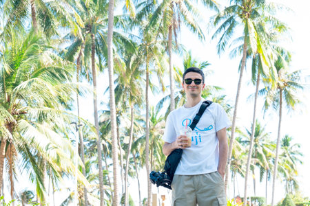 Young Male Traveler with Iced Coffee and Camera Gear Under Tropical Palm Trees, Bright Sunny High-Key Lighting Portrait with Copyspace for Travel Blogs Nha Trang 20.12.2025 Vietnamのeditorial素材
