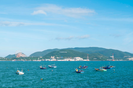 Small colorful fishing boats float on blue water with a distant coastal resort town, a Ferris wheel, and a green mountain under a clear sunny sky now. Nha Trang 20.12.2025 Vietnamのeditorial素材
