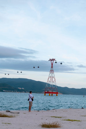 A man in a white t-shirt and shorts stands on a sandy beach looking towards a large red cable car tower situated in the blue ocean under a cloudy sky. Nha Trang 20.12.2025 Vietnamのeditorial素材