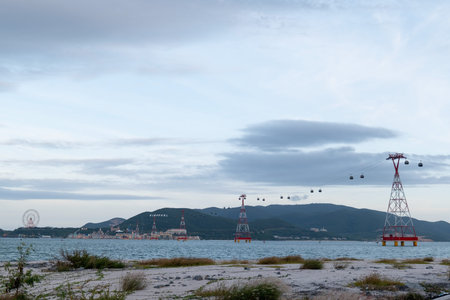 A coastal landscape featuring red and white cable car towers in the ocean, a sandy foreground with bushes, and green mountains under an overcast sky.. Nha Trang 20.12.2025 Vietnamのeditorial素材
