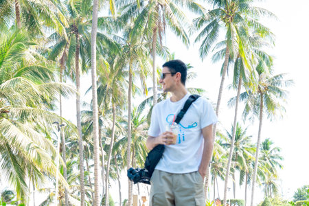 Young Male Traveler Relaxing in Tropical Palm Grove with Iced Coffee; Authentic Summer Vacation Lifestyle Shot with High-Key Cinematic Sun Lighting Nha Trang 20.12.2025 Vietnamのeditorial素材