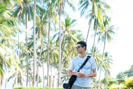 Young Male Digital Nomad Traveler with Sunglasses and Iced Coffee Standing Under Tropical Palm Trees, Bright Natural Sunlight, Cinematic Outdoor Lifestyle Portrait Nha Trang 20.12.2025 Vietnamのeditorial素材