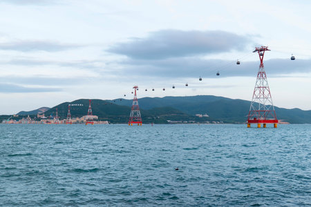 Overhead cable car system features red and white metal pylons over a body of water with mountains and coastal buildings under the grey sky at daytime. Nha Trang 20.12.2025 Vietnamのeditorial素材