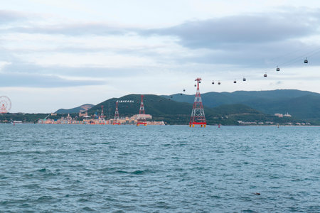 Aerial cable car system with red and white lattice towers spans over blue sea water towards a coastal resort with hillside signage and a Ferris wheel. Nha Trang 20.12.2025 Vietnamのeditorial素材