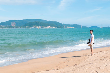 Solo Young Male Traveler Standing on Sunny Tropical Beach with Vinpearl Nha Trang Skyline Background, Bright Coastal Tourism Lifestyle with Copy Space Nha Trang 20.12.2025 Vietnamのeditorial素材