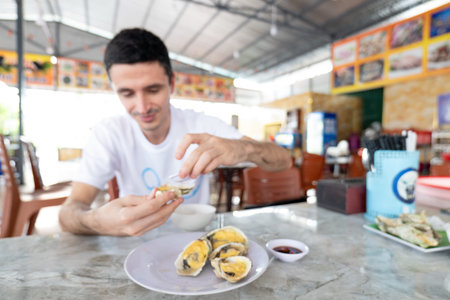 A Caucasian man in a white shirt sits at marble table, handling grilled oysters with a yellow topping served on a plate in an open-air restaurant area. Nha Trang 20.12.2025 Vietnamのeditorial素材
