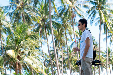 Adult male wearing white T-shirt and sunglasses stands in profile holding a plastic cup with a straw against dense green tropical coconut palm trees. Nha Trang 20.12.2025 Vietnamのeditorial素材