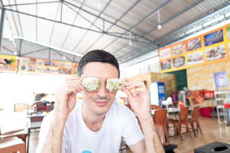 Adult Caucasian male wearing a white t-shirt holds two closed oyster shells over his eyes while sitting at a table within a semi-outdoor dining space. Nha Trang 20.12.2025 Vietnamのeditorial素材
