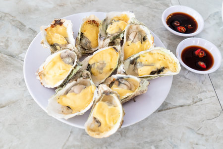 A white plate holds the grilled oysters in shells topped with melted yellow cheese sauce alongside two small bowls of soy sauce and red chili slices.の写真素材