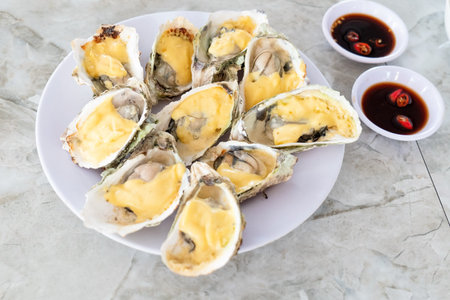 Ten baked oysters in shells with yellow cheese on a white plate. Two bowls of dark sauce with chili are next to the plate on a stone pattern tabletop. Vietnamの写真素材